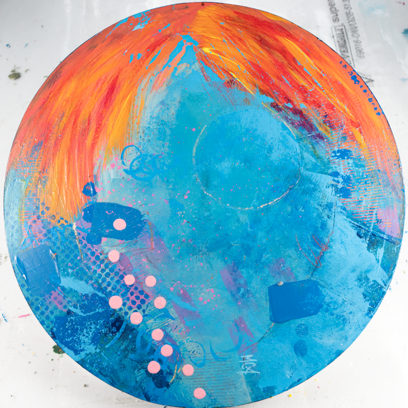 Close-up of an abstract circular painting. The dominant colors are vibrant orange and turquoise blue, blended with streaks of yellow, pink, and purple. The texture appears layered and brushstroked, creating a dynamic visual effect.