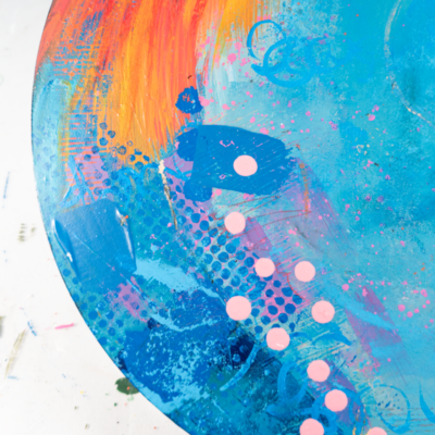 Close-up of an abstract circular painting. The dominant colors are vibrant orange and turquoise blue, blended with streaks of yellow, pink, and purple. The texture appears layered and brushstroked, creating a dynamic visual effect.