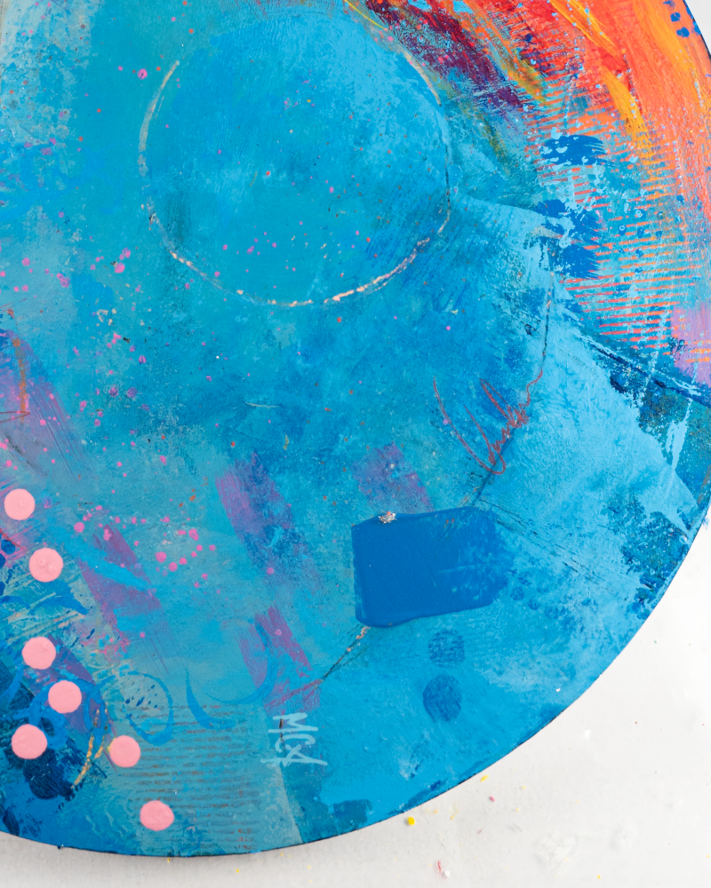 Close-up of an abstract circular painting. The dominant colors are vibrant orange and turquoise blue, blended with streaks of yellow, pink, and purple. The texture appears layered and brushstroked, creating a dynamic visual effect.